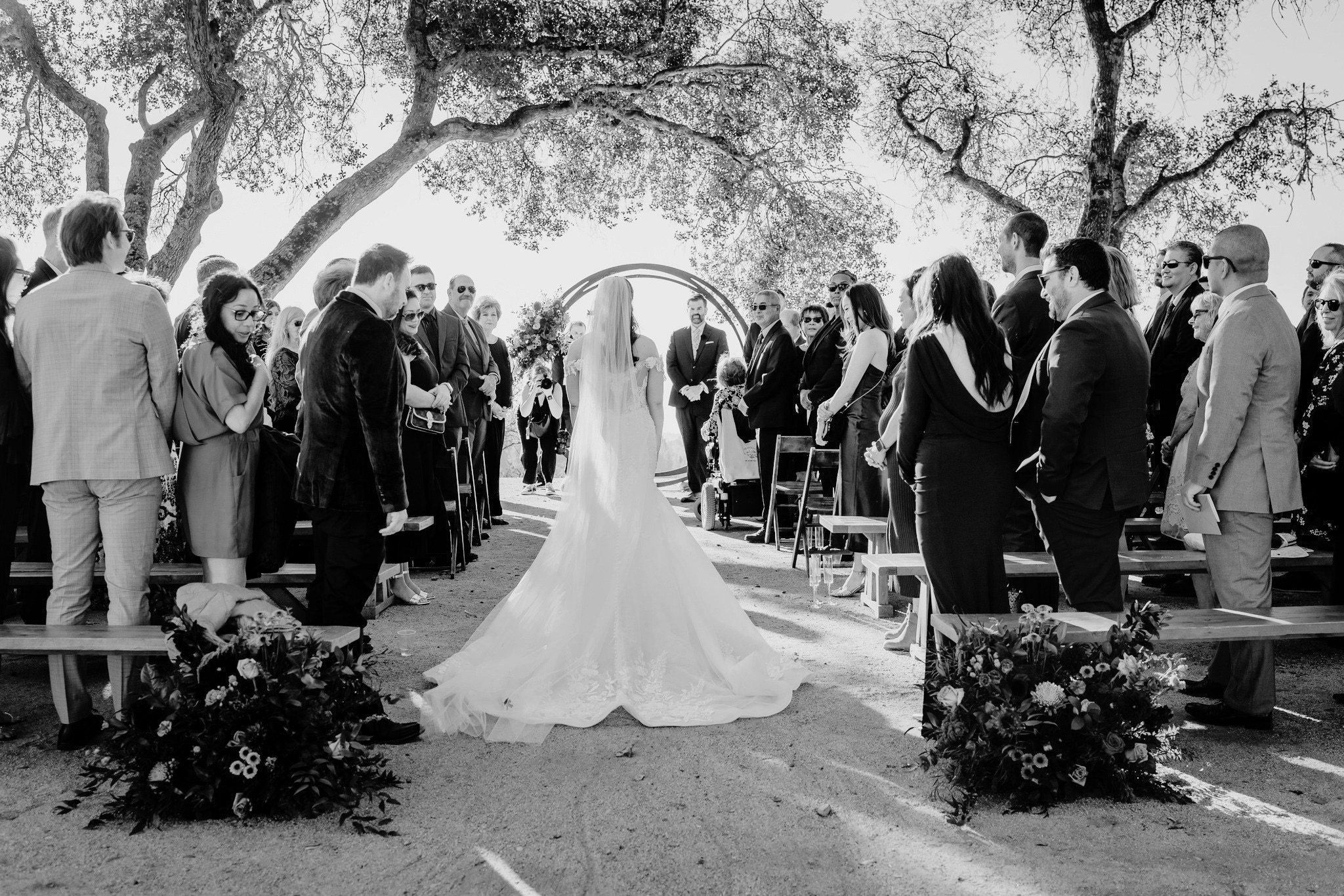 A bride and groom walk hand in hand with their wedding party across a coastal lawn